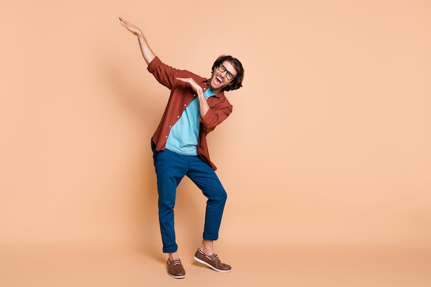 Photo full length photo of guy dance raise hands have fun wear spectacles brown t-shirt trousers shoes isolated beige color background