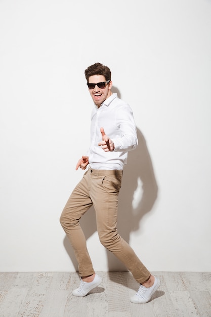 Full length photo of handsome man wearing black sunglasses in stylish clothing dancing and pointing index finger on camera, isolated over white space with shadow