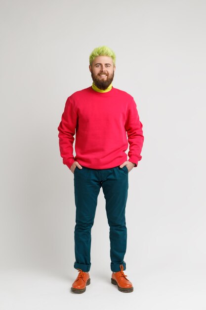 Full length photo of happy smiling bearded man with bright appearance wearing red sweater pants and shoes keeps hands in pockets Indoor studio shot isolated on gray background