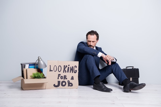 Full length photo of  loser worker mature guy jobless handmade placard search work sit floor briefcase belongings box lost success career crash wear suit shoes isolated grey background