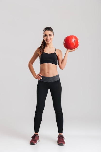 Full-length photo of pleased sportswoman looking  and holding fitness ball, isolated over gray wall