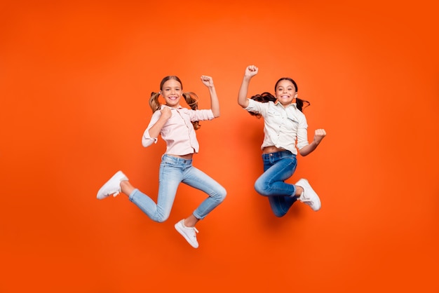 Full length photo of positive cute two children girls jump celebrate victory in school contest raise fists show strength wear white shirt denim jeans isolated orange color background