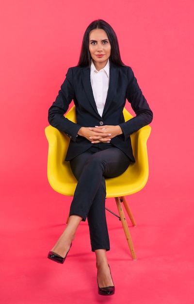 Full-length photo of a serious woman in a black suit, who is posing while sitting in her yellow armchair with her right hand on her knee and looking piercingly in the camera