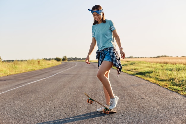 Foto a figura intera di una donna snella e attraente che fa acrobazie su uno skateboard, si diverte da sola per strada, cavalca un longboard, indossa abiti casual, guarda in basso.