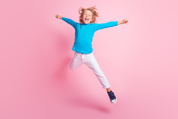 작은 소년 점프 스타의 전체 길이 사진은 파란색 터틀넥 바지 운동화를 신고 파스텔 핑크색 배경에서 분리되었습니다.