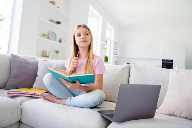 Full length photo of smart pensive geek nerd kid girl sit divan legs crossed write home task copybook think thoughts pencil touch chin in house indoors