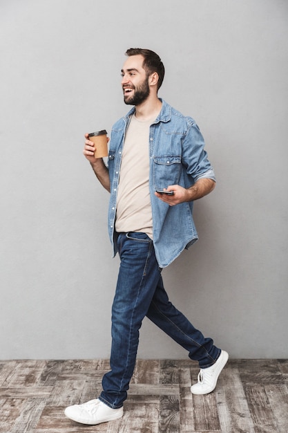 Full length photo of smiling european man having beard using smartphone and drinking takeaway coffee isolated over gray wall