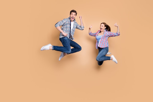 full length photo of two people crazy lady guy jumping high raising thumbs up celebrating successful win wear casual plaid jeans clothes isolated beige color background