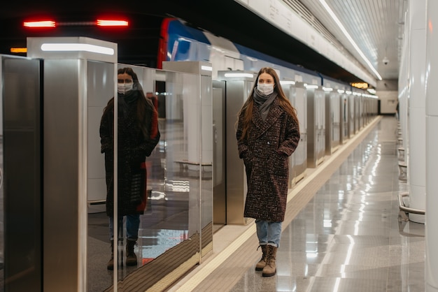 地下鉄のホームで出発する電車の近くに立っているコロナウイルスの蔓延を防ぐために、医療用フェイスマスクを着用した女性の全身写真。マスクをした女の子が社会的距離を保っています。