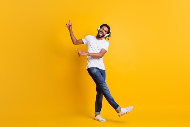 Foto a figura intera di un giovane ragazzo di colore diretto con il dito in alto a bocca aperta indossa le cuffie t-shirt bianca jeans scarpe da ginnastica isolate sfondo di colore giallo