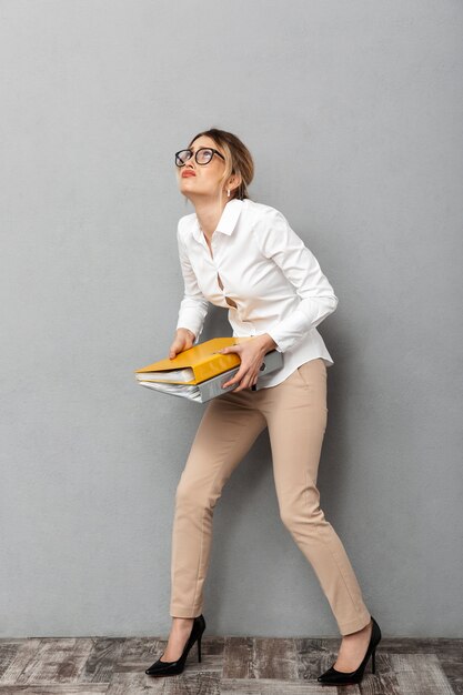 Full length photo of young businesswoman wearing glasses holding paper folders in the office, isolated 