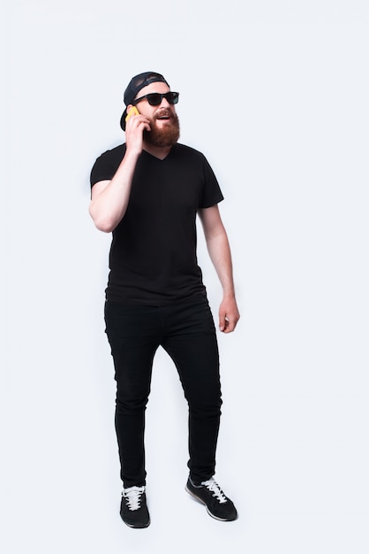 Full length photo of young guy with beard talking on smartphone