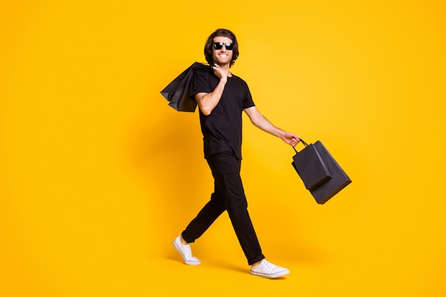 Full length photo of young man walk bear mall bags wear black t-shirt pants white sneakers sunglass isolated yellow color background