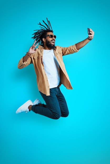 full length of playful young man in casual wear taking selfie and smiling while hovering against blue wall