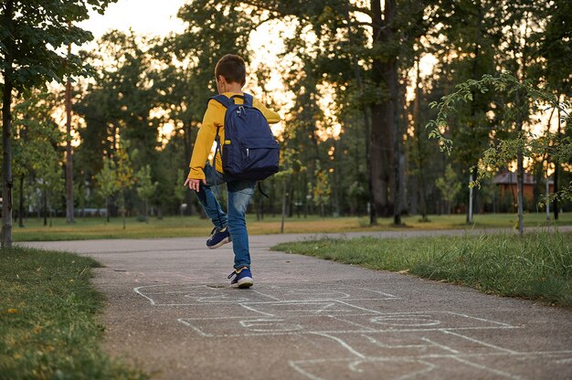 Full-length portrait of an active schoolboy enjoying recreation, playing hopscotch on the ground after first day at school. Street children's games in classics.