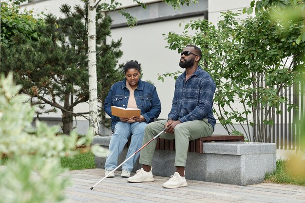 Photo full length portrait of adult couple with partner with visual disability sitting on bench in park to