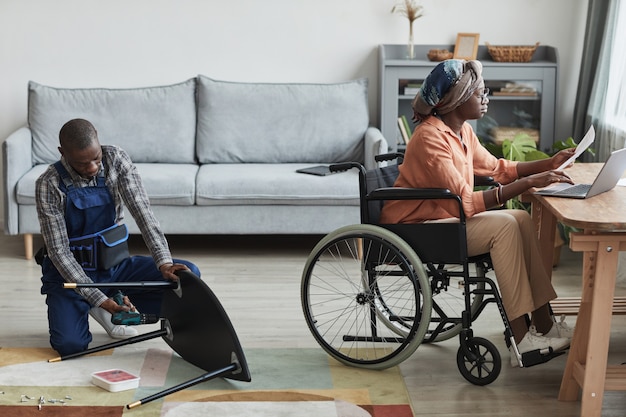 Ritratto a figura intera di uomo afro-americano che assembla mobili all'interno della casa con donna in sedia a rotelle che lavora alla scrivania, servizio tuttofare e concetto di assistenza, spazio copia