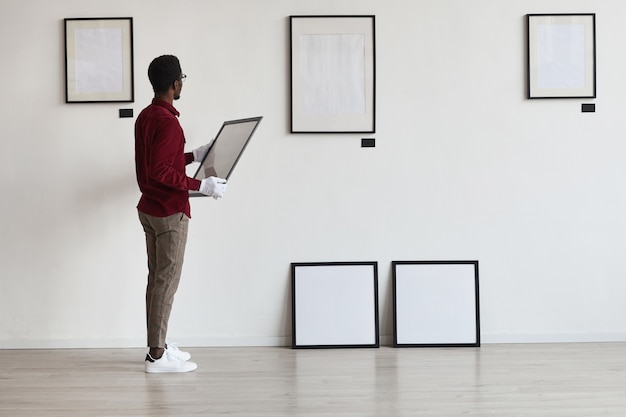 Full length portrait of African-American man planning art gallery or exhibition while setting up frames on white wall, 