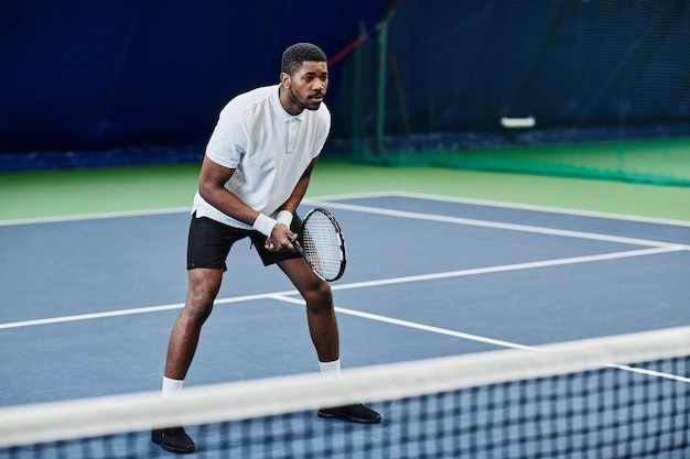 Full length portrait of agile black sportsman playing tennis at indoor court copy space