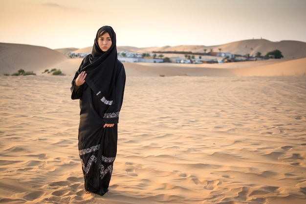 full length portrait of arabic woman in traditional black clothes standing in the desert in front of bedouin village