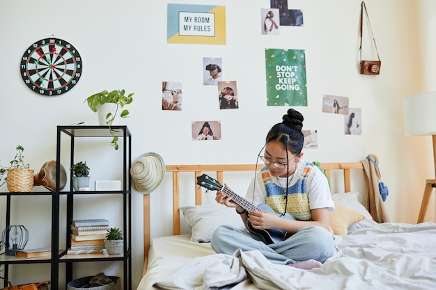 Full length portrait of Asian teenage girl playing ukulele while sitting on bed in cozy room, copy space