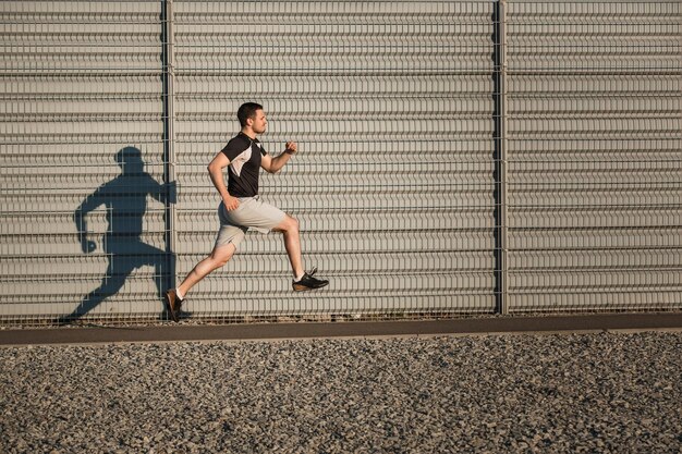 Full length portrait of athletic man running along beautiful grey wall outdoors, muscular build young runner working out while jogging in the park. Sunset