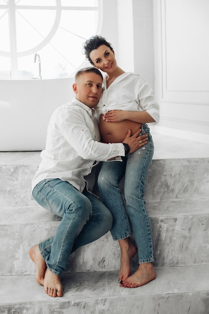 Full length portrait of attractive adult Caucasian couple in white shirts and jeans posing barefoot on grey steps and looking at camera. Husband embracing pregnant wife.