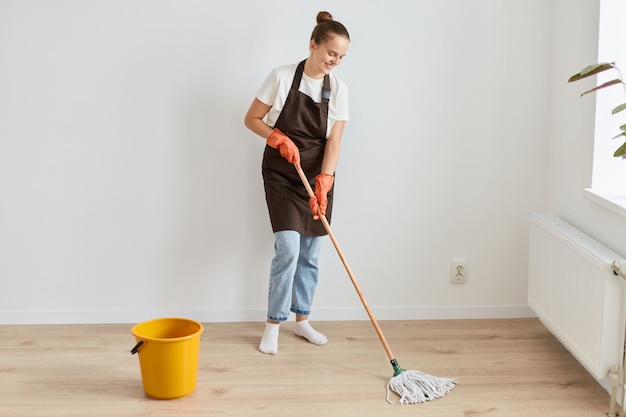 Ritratto a figura intera di attraente donna snella che indossa maglietta bianca e grembiule in posa con attrezzature per la pulizia e lavaggio del pavimento nel suo soggiorno facendo le faccende domestiche