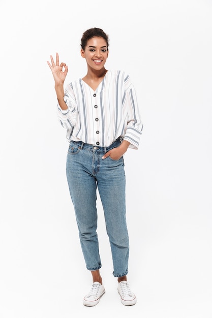 Full length portrait of an attractive young african woman standing isolated over white wall, showing ok