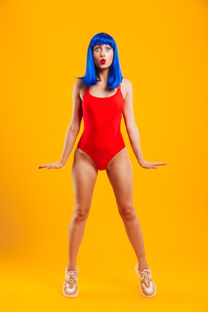 Full length portrait of an attractive young funky girl with blue hair wearing swimsuit standing isolated over yellow wall, posing