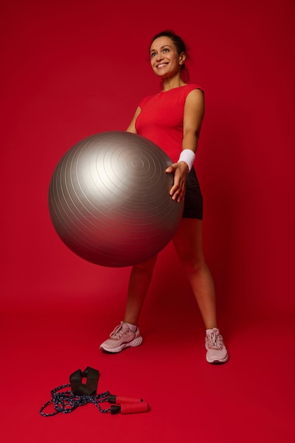 Full length portrait of a beautiful active woman in sportswear exercising with fit ball against red colored background with copy space