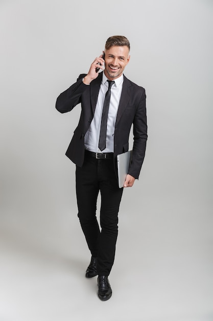 Full length portrait of beautiful adult businessman in office suit holding laptop and talking on smartphone while standing isolated