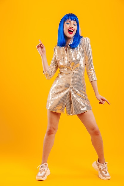 Full length portrait of a beautiful happy young woman with blue hair wearing shiny dress standing isolated over yellow wall, dancing
