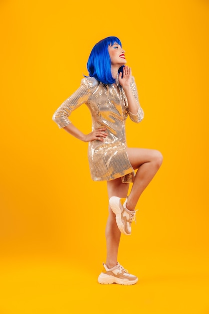 Full length portrait of a beautiful smiling young woman with blue hair wearing shiny dress standing isolated over yellow wall, posing