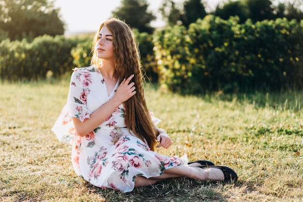 Full length portrait of beautiful woman with long curly hair on currant field. woman in a light dress sits on the grass in sunny day