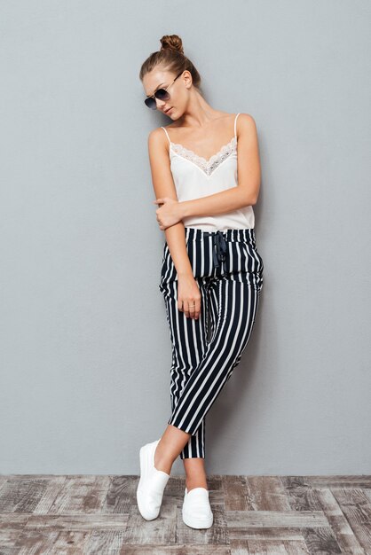 Full length portrait of a beautiful young girl in sunglasses posing and looking away isolated on the gray background