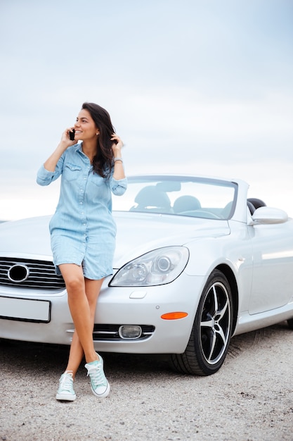 Full length portrait of a beautiful young woman talking on smartphone and standing at her car