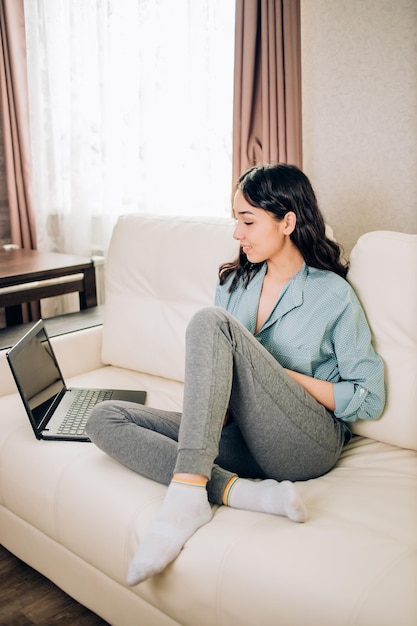 Full length portrait of brunette sitting with laptop computer, laughing watching funny video from social networks. Pretty woman with positive smile relaxed in spacious white room in front of windows.