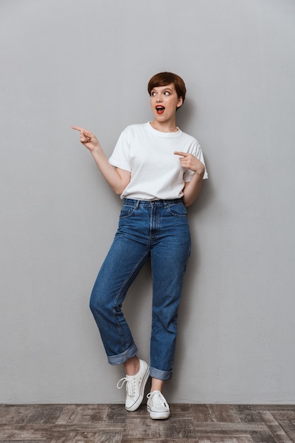 Full length Portrait of caucasian brunette woman wearing jeans pointing finger aside at copyspace isolated over gray wall