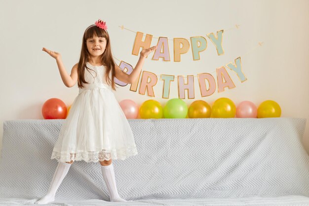 Full length portrait of charming dark haired little girl standing on sofa wearing white dress posing with balloons and party inscription on background standing with raised arms