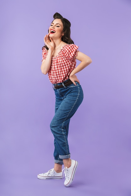 Full length portrait of charming pin-up girl in american style wear smiling at camera isolated over violet wall