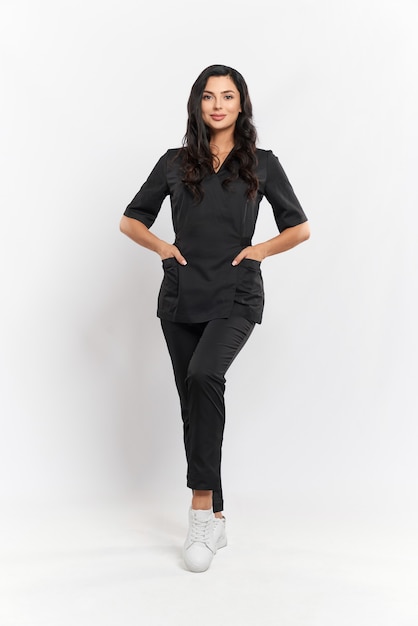 Full length portrait of charming professional beautician in black medical uniform standing on white