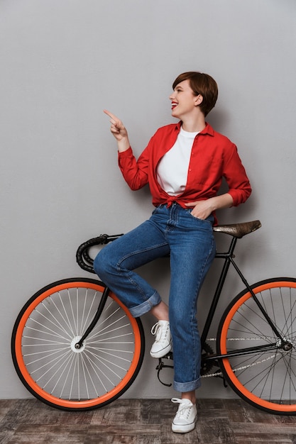 Full length Portrait of charming woman pointing finger aside and standing by bicycle isolated over gray wall