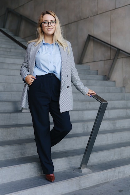 Full length portrait of charming woman in stylish office\
clothing standing on steps of office building. business lady with\
blond hair smiling and looking aside.