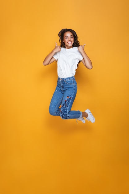 Full length portrait of a cheerful girl with long dark hair jumping over yellow wall, showing thumbs up