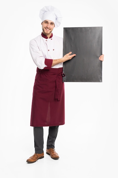 白い背景で隔離の空白のボードを保持している制服を着た陽気な男性シェフ料理人の完全な長さの肖像画。