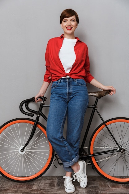 Full length Portrait of cheerful woman smiling and standing by stylish bicycle isolated over gray wall