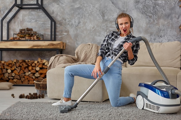 Full length portrait of cheerful young woman 20s listening to music via headphones and having fun with vacuum cleaner in the house.