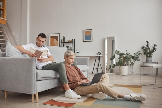 Полнометражный портрет современной гей-пары, использующей компьютеры, во время совместного отдыха на диване в минималистичном домашнем интерьере, копировальное пространство
