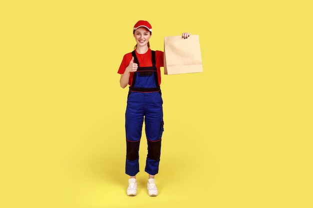 Full length portrait of courier woman holding pointing at ordered parcel, delivering food in paper bag, post mail services, wearing overalls and cap. Indoor studio shot isolated on yellow background.